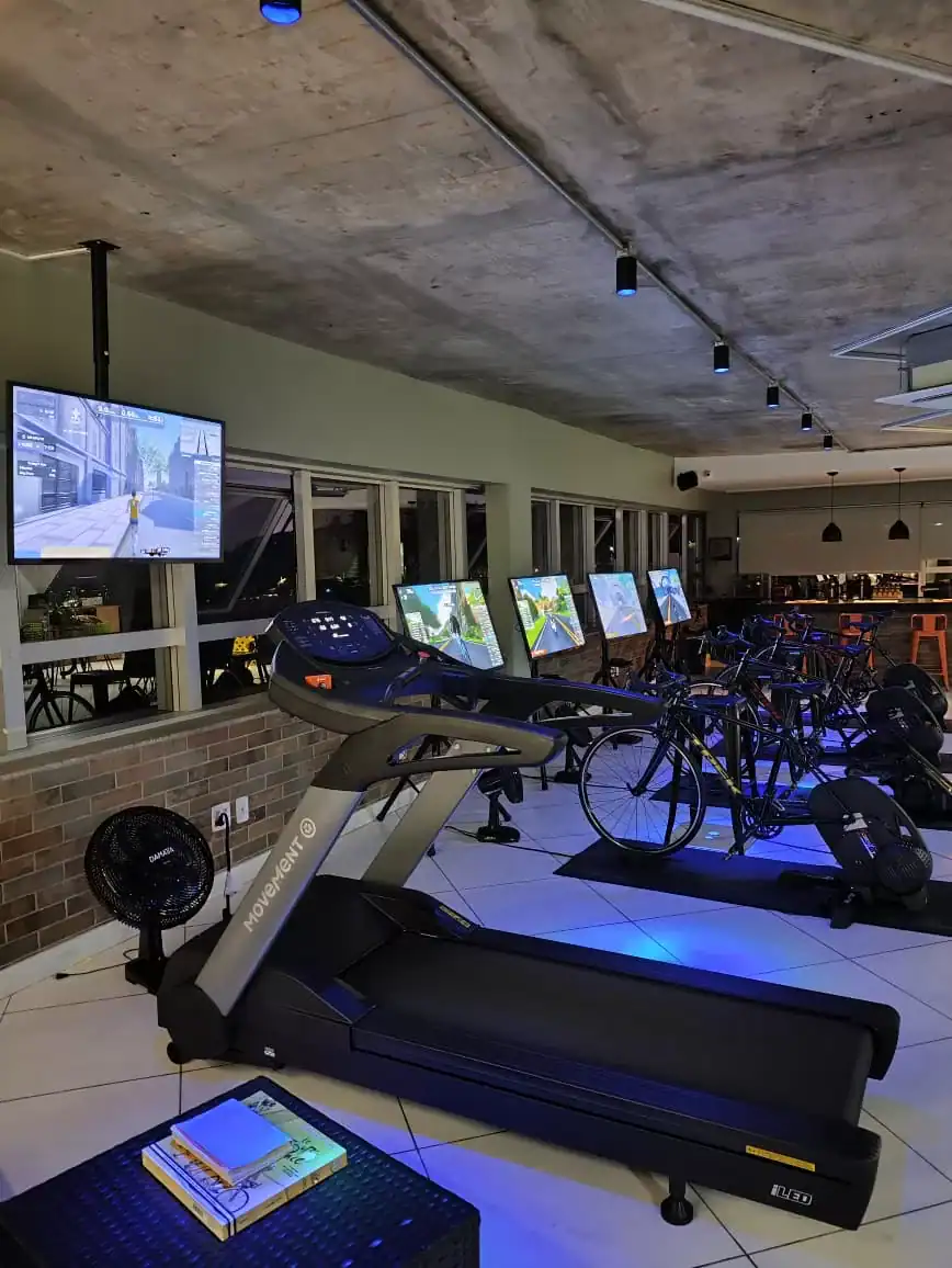 Alto texto: Este ambiente de treino com esteiras, bicicletas ergométricas e telas interativas da Velotime Training® oferece uma experiência avançada de treinamento indoor, promovendo saúde e performance.